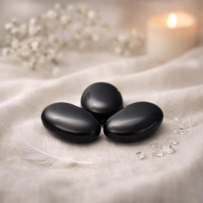 Schwarzer Obsidian als Handschmeichler auf hellem Stoffhintergrund mit kerze und weißer Blüte als Symbol für die Schutzsteine Übersicht.